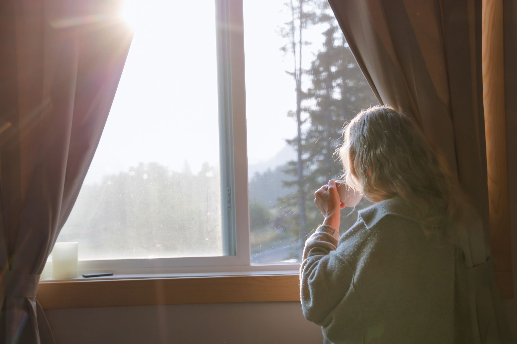 Woman enjoying a hot beverage while looking out a window at sunrise over trees